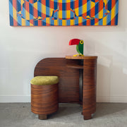 Art Deco Walnut vanity with a green and red parrot figurine, next to a round stool with a green cushion, against a white wall with colorful abstract art.